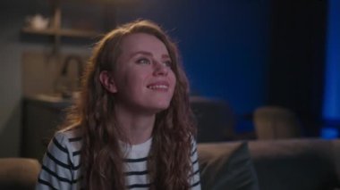 Şirin sarışın kıvırcık kadın televizyonu açıyor, akıllı TV uygulamalarında geziniyor ve evdeki oturma odasında kanepede otururken kendini eğlendirmek için kanalları seçiyor. Sinematik çekim.