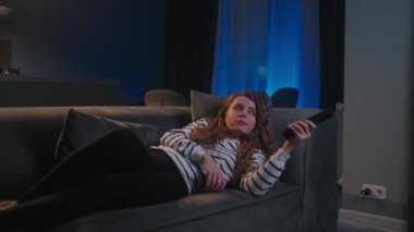 Şirin sarışın kıvırcık kadın televizyonu açıyor, akıllı TV uygulamalarında yol gösteriyor ve evdeki oturma odasındaki kanepede uzanmış kendini eğlendirmek için kanalları seçiyor. Sinematik bir çekim..