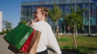 Alışveriş merkezinden şehre elinde poşetlerle gelen mutlu kadın. Heyecanlı kız, Kara Cuma indiriminden zevk alıyor. Dişi danslar ve kameraya poz vermek. Yavaş çekimde arka plan.
