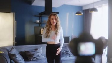 Sosyal medya platformunda beğeniler ve takipçiler için moda dans yarışması videosu kaydeden neşeli kız. Komik dans eden bir kadın, ev stüdyosundaki oturma odasında gösterisini kameraya çekiyor..