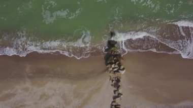 Havadan aşağı sakin, boş bir sahil manzarası, kıyıda kum yıkayan köpüklü okyanus dalgaları. Deniz dalgaları taştan dalgalanmalara ve kumlu kaplamalara çarpıyor. Yukarıdan görüntüle, İHA sağa hareket eder.