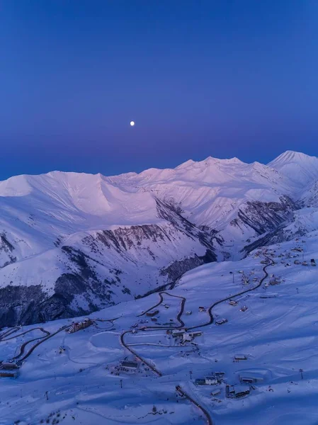 Aerial of snowy mountain range on winter sunrise at ski resort. Moon above mountains valley and village with switchbacks road at a sunset. Caucasus peaks skyline in the dusk. City lights at night.