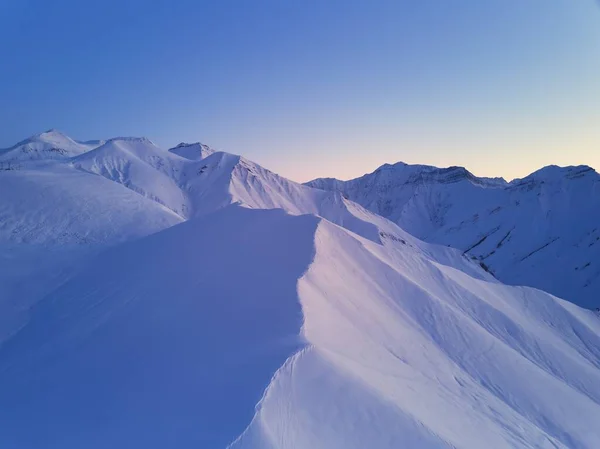 Aerial of snowy mountain slopes full of powder for freeride at ski resort on winter sunrise. Mountains range of backcountry covered with virgin snow. Caucasus peaks skyline with a twilight afterglow.