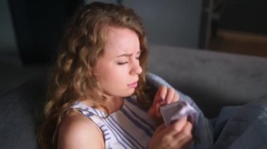 Soğuk algınlığı olan bir kadın evde ateşini kontrol etmek için battaniyenin altında öksürüyor ve burnunu siliyor. Beyaz kadın koronavirüsten muzdarip. 19 derece izole edilmiş. Termometreye bakıyor.