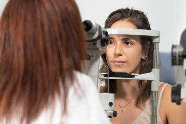 Optometrist bir kadın, başka bir kadının gözlerini kesikli lambayla inceliyor.