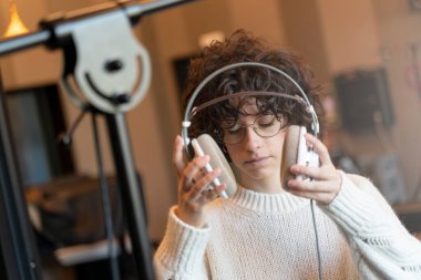 Genç, kıvırcık saçlı, gözlüklü bir kadın gerçek bir stüdyoda şarkı kaydediyor. 