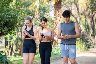 Üç genç sporcu parkta telefonlarına yoğunlaşarak yürüyor.