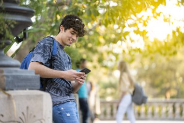 Genç bir öğrenci telefonuna bakıyor ve sokakta gülümsüyor.