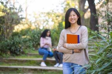 Genç Asyalı kız öğrenci gülümsüyor ve elinde bir defterle kameraya bakıyor.