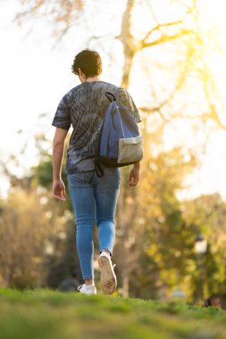 Çantalı genç adam arka planda ağaçlarla parkta dolaşıyor.