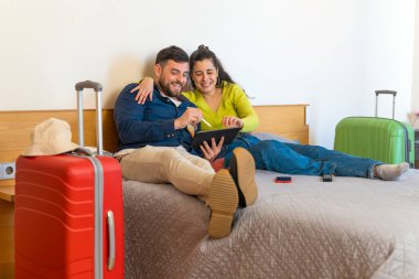 Genç turist çift oteldeki yatağın üzerine tablet çiziyor.