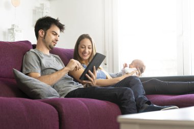 Anne ve baba yeni doğmuş bebeklerini dijital tabletle kanepede oturtuyorlar.