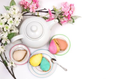 Beyaz bir arkaplanda, beyaz bir çaydanlık etrafında, beyaz bir çaydanlık etrafında servis edilen çok renkli pastalar. Çiçek açan dallar, düz bir zemin, beyazda izole edilmiş, sağda kopyalanmış, yatay.