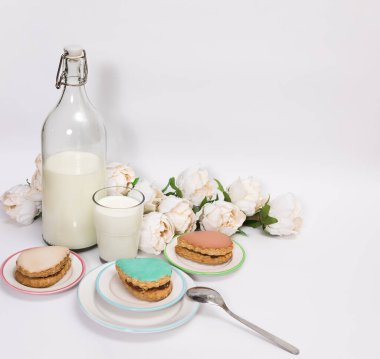 Çok renkli kekler (turkuaz, bej, karamel) beyaz arka planda bir şişe ve bir bardak sütle çevrili çay tabakları üzerinde servis edilir.