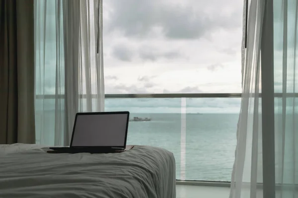 Workation concept. Empty screen laptop on bed and ocean view balcony.