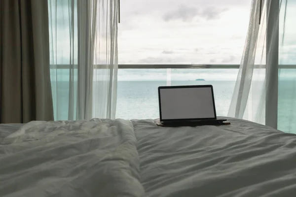 Workation concept. Empty screen laptop on bed and ocean view balcony.