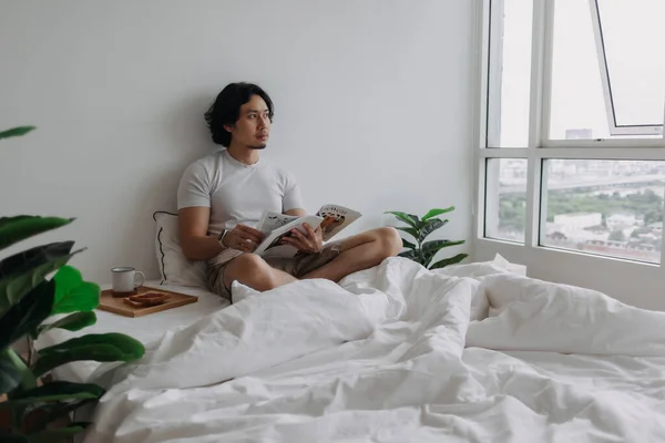 Hombre asiático leyendo libro en la cama con panes y café en el apartamento. —  Fotos de Stock