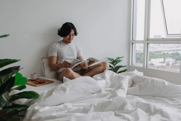 Hombre asiático leyendo libro en la cama con panes y café en el apartamento. —  Fotos de Stock
