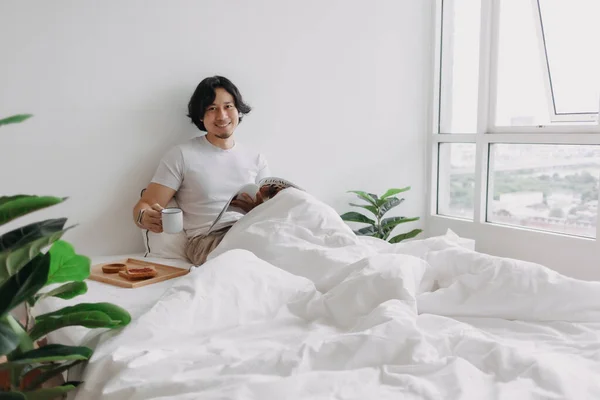 Homem asiático lendo livro na cama com pães e café no apartamento. — Fotografia de Stock