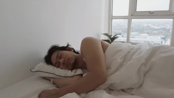 El hombre se despierta en la cama blanca en su apartamento de dormitorio en una acogedora mañana perezosa. — Vídeos de Stock