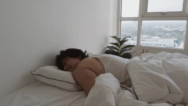 Seorang pria bangun di tempat tidur putih di apartemennya di pagi yang nyaman dan malas. — Stok Video