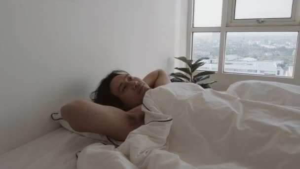 El hombre se despierta en la cama blanca en su apartamento de dormitorio en una acogedora mañana perezosa. — Vídeo de stock