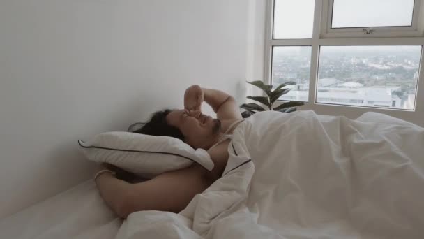 Homem acorda na cama branca em seu apartamento de quarto em uma manhã preguiçosa aconchegante. — Vídeo de Stock