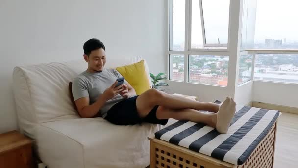 Человек смотрит на свой мобильный телефон, расслабляясь на диване в своей гостиной. — стоковое видео