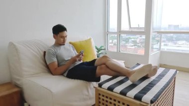 Asyalı adam oturma odasındaki kanepede dinlenirken cep telefonuna bakıyor..