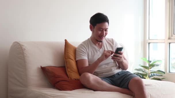 幸せなアジアの男はカジュアルな日にリビングルームで彼の携帯電話を使用します — ストック動画