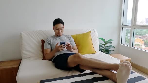 アジアの男は彼のソファの上でリラックスしながら モバイルアプリケーションに満足 — ストック動画