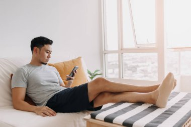 Asian man looking at his mobile while relax on sofa in his living room.