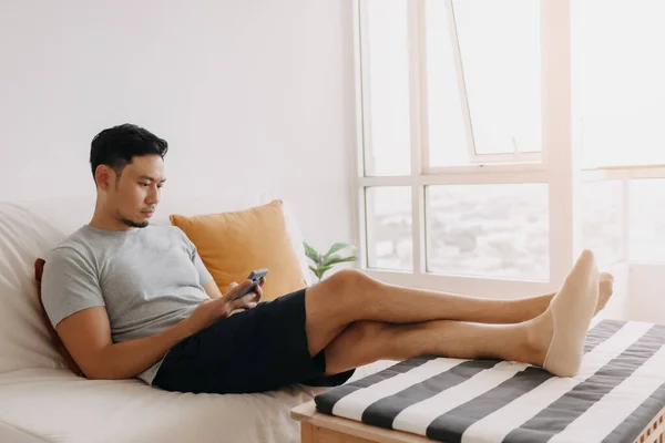 Asian man looking at his mobile while relax on sofa in his living room.