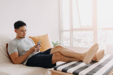 Asian man looking at his mobile while relax on sofa in his living room.
