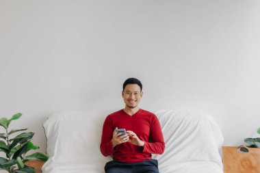 Happy asian man on sofa use phone for trading business with advertising space. 