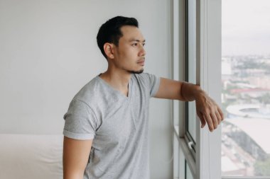 Thinking serious lonely asian man stand lean on the window in his apartment