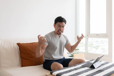 Angry and furious Asian trader man working on tablet or laptop in his apartment.