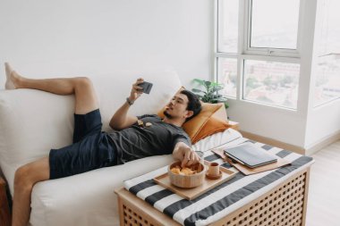 Asian man watch online tv and eating snack instead of working from home.