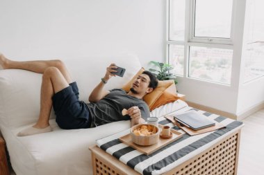 Asian man watch online tv and eating snack instead of working from home.