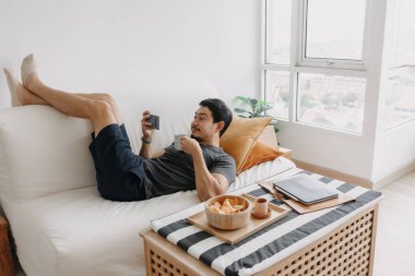 Asian man watch online tv and eating snack instead of working from home.