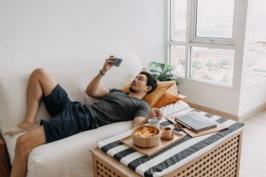 Asian man watch online tv and eating snack instead of working from home.
