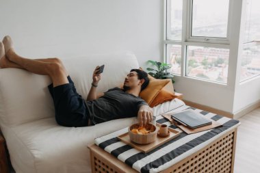 Asian man watch online tv and eating snack instead of working from home.