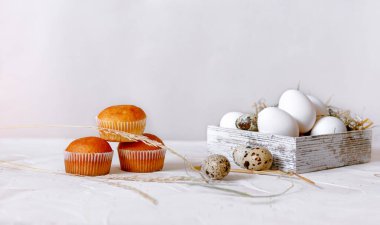 Paskalya 'da hayat. Beyaz tavuk yumurtaları ve bıldırcın yumurtaları ahşap bir kutuda, arka planda saman ve beyaz masa yüzeyinde kapkekler var.