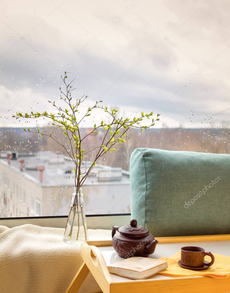 Spring twigs in the bottle on the table with a plaid, pillow, book, teapot and a cup with a rainy city outside of the window (on the background). Concept of a feeling of coziness at home.
