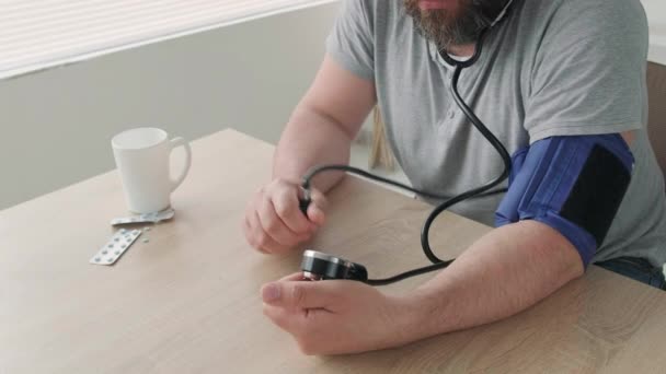 男は窓のそばのテーブルに座っている間 血圧計で血圧を測定します 薬とマグカップがテーブルの上にある — ストック動画