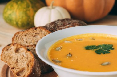 Vejetaryen sonbahar balkabağı çorbası ve ev yapımı ekmek. Beyaz çorba kasesi maydanozla süslenmiş, kabak çekirdeği ve susam tohumları kabakların arka planında.