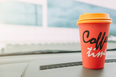 Arabada kahve var. Araba konsolunda parlak turuncu kahve fincanı (panel). Kahve saatine yazılmış.