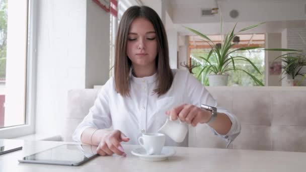 Девушка в белой рубашке, сидящая в кафе, наливает молоко в кофе. — стоковое видео
