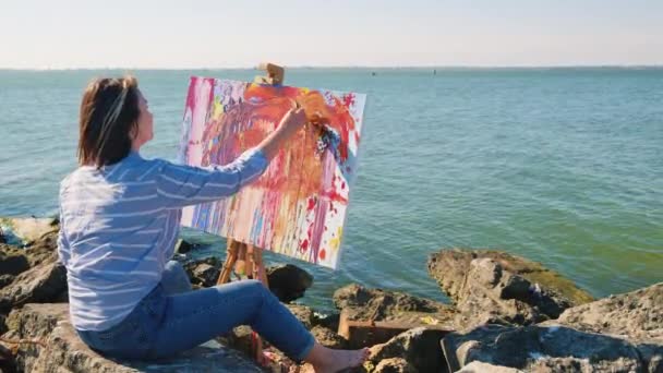 Talentierte Künstlerin sitzt auf Steinen und malt mit einem Spachtel ein Bild auf Leinwand am Flussufer. — Stockvideo