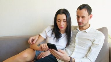 Uygulamada akıllı telefona bakan ya da satın almak için ürün seçen bir çift. Evdeki kanepede oturmak. Kız, erkeğin telefon ekranından seçmesine yardım ediyor. Adam telefona bakarken tatmin oluyor.